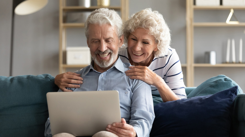 Happy elderly 50s husband and wife relax on comfy couch in living room watch funny video on laptop together