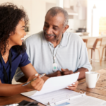 Smiling female caregiver in scrubs looks over older adult woman's shoulder at tablet