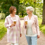 Two older adult women wearing jackets walking through park with coffee cups