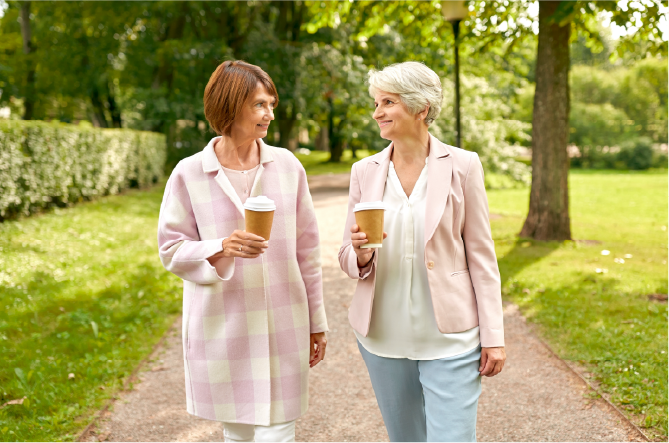 Two older adult women wearing jackets walking through park with coffee cups